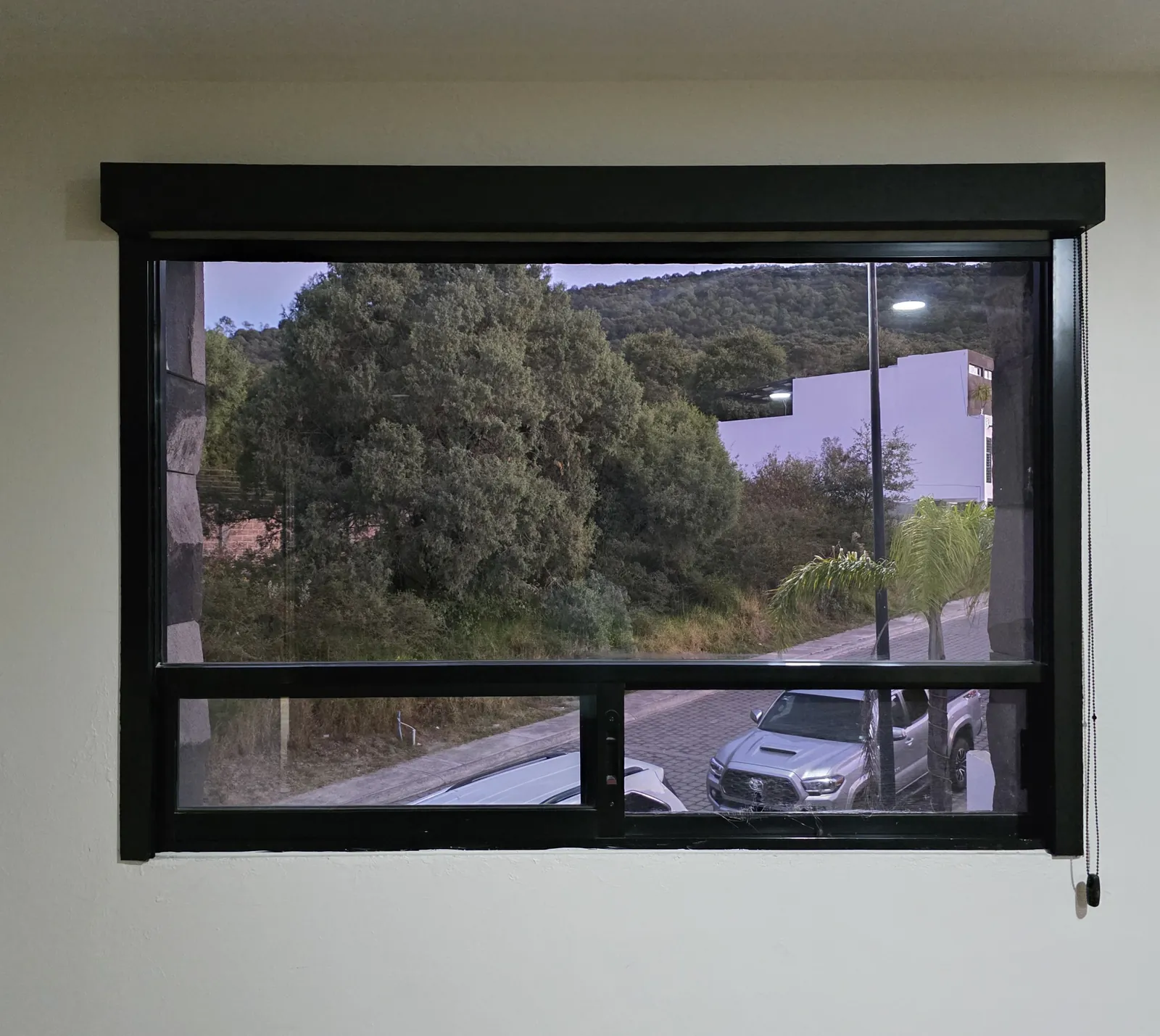 Ventana real con Sistema Fit de Style Coverings abierto: cajón negro, guías laterales y vista a los árboles y la calle